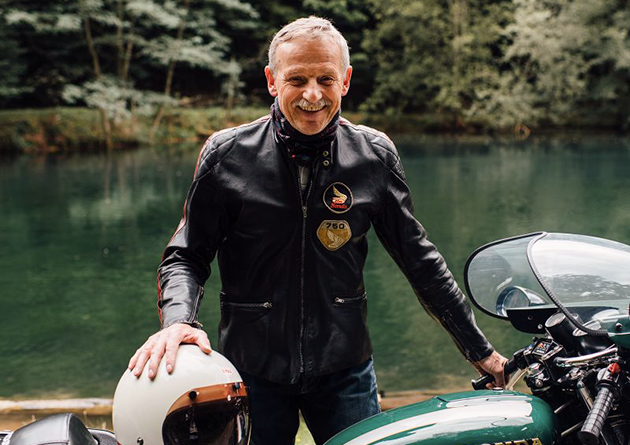 El fundador y director general de Hepco & Becker está delante de un lago con su motocicleta y sonríe a la cámara.