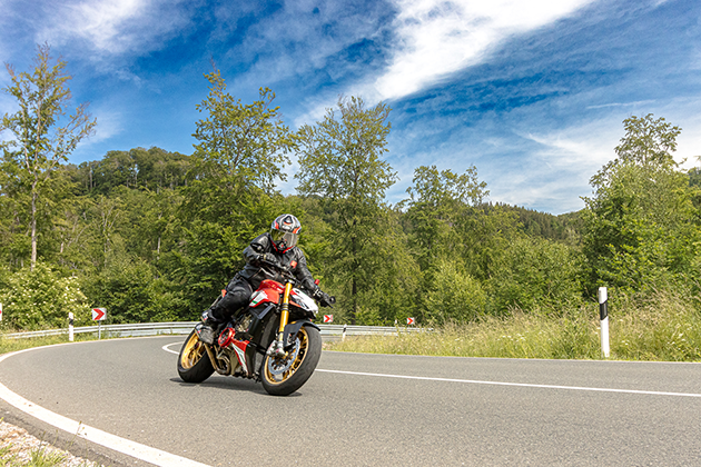 A motorcyclist in black clothing and helmet rides a red, converted Ducati Streetfighter along a winding mountain road in Germany.