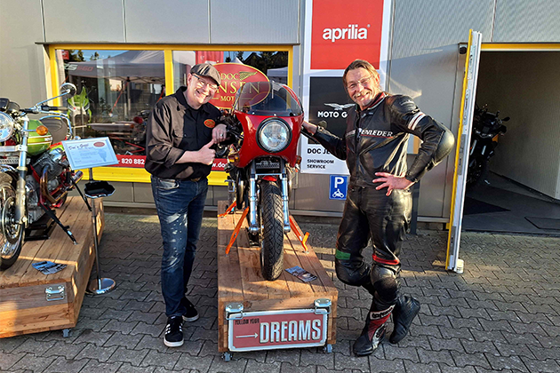 Jens Koch y su cliente están de pie a izquierda y derecha de una Moto Guzzi roja, más vieja, delante de la tienda de Doc Jensen. Ambos sonríen. El cliente lleva un traje de cuero negro.