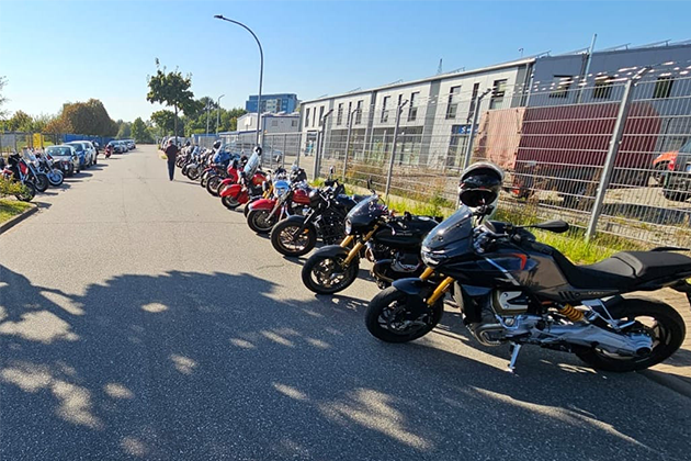 Varios modelos diferentes de motocicletas están aparcados al borde de la carretera. Al fondo se ve un polígono industrial. El sol brilla bajo un cielo azul.