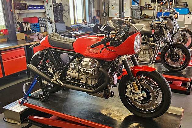 Varios modelos antiguos de Moto Guzzi reconstruidos están sobre plataformas elevadoras rojas en el taller de Doc Jensen. La Guzzi en primer plano es roja.