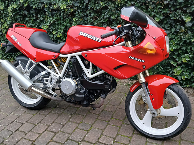 A red 1991 Ducati 750SS is parked on a paved path in front of a hedge.