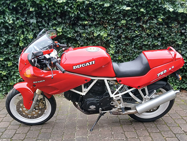 A red 1991 Ducati 750SS is parked on a paved path in front of a hedge.