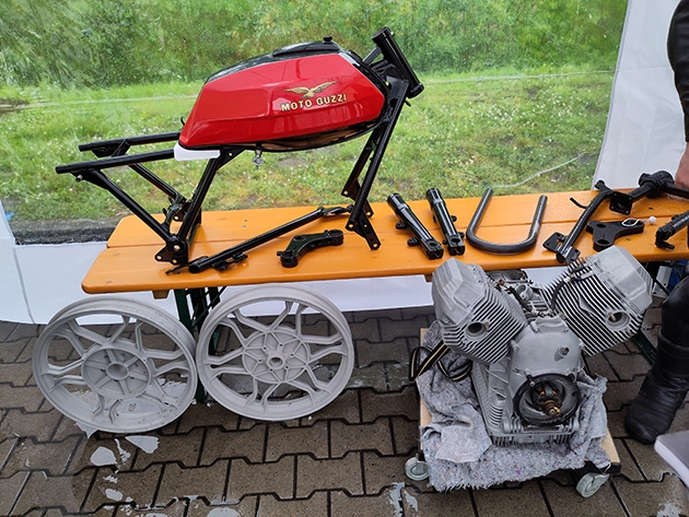 You can see a red petrol tank with the Moto Guzzi logo, two silver wheel rims, a silver V2 engine block and various frame and suspension parts.