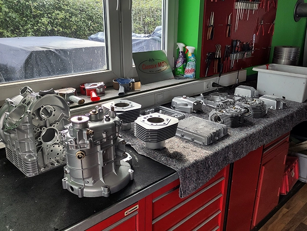 Several freshly restored Moto Guzzi engine parts are lying on a workbench. The parts include cylinder heads, cylinder blocks, valve covers and other components in perfect condition.