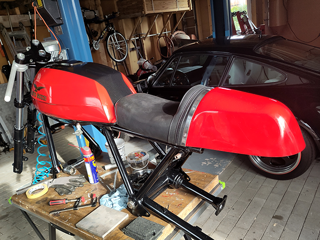 You can see a red petrol tank with the Moto Guzzi logo, two silver wheel rims, a silver V2 engine block and various frame and suspension parts.