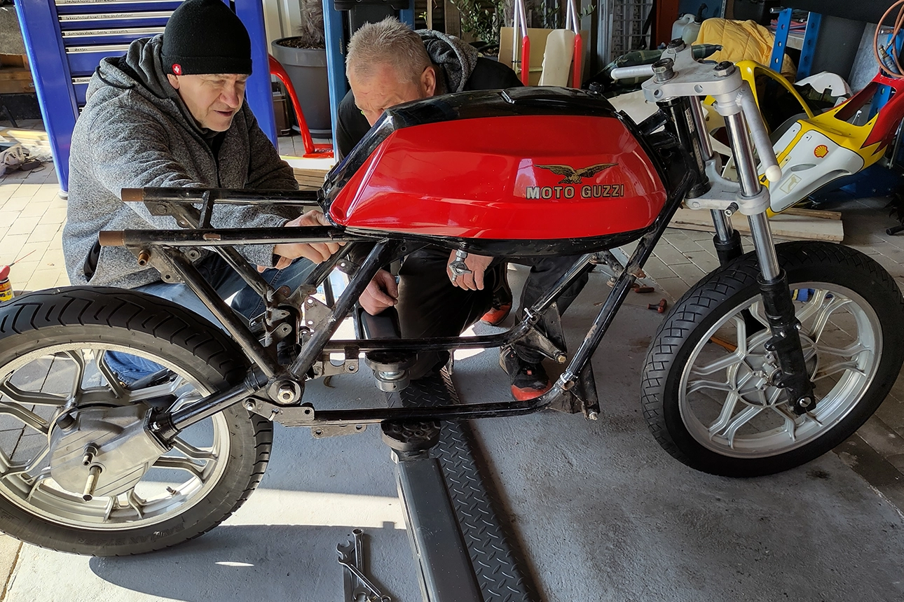 Two men work on a partially dismantled Moto Guzzi 850 T5 P Caferacer from 1983 with a red tank. Tools and motorcycle parts are in the background.