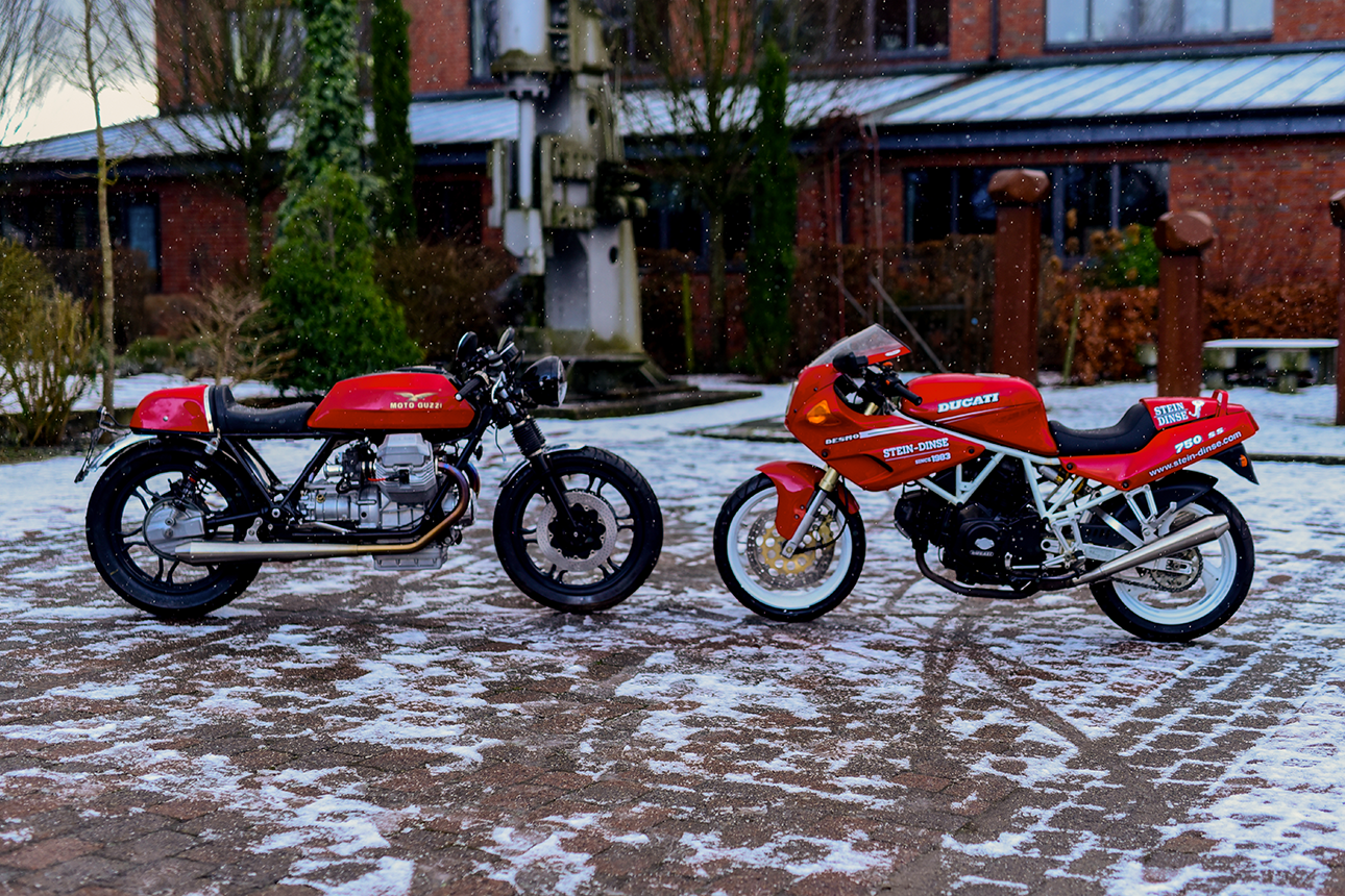 Two men work on a partially dismantled Moto Guzzi 850 T5 P Caferacer from 1983 with a red tank. Tools and motorcycle parts are in the background.